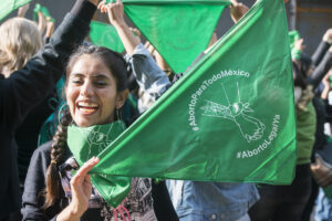 Es la fotografía de una mujer joven que con sus manos sostiene frente a ella un pañuelo verde, símbolo de la lucha feminista. Detrás de ella aparecen otras mujeres de espaldas también con pañuelos.