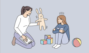 Imágen de psicóloga en sus rodillas, tomando un conejo de peluche y mostrándoselo a una niña sentada en el piso que se sostiene las rodillas. 