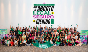 Fotografía en plano general que retrata a varias personas que acudieron al Encuentro Nacional por el Aborto Legal y Seguro en México. A todas se les ve sentadas. Algunas alzan el puño y sujetan un pañuelo verde, mientras que otras sonríen y gritan consignas.