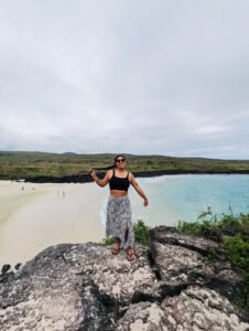 Fotografía en plano general de Estefania. Ella es una mujer joven de piel morena y cabello largo color negro. Luce sonriente sobre unas rocas en Puerto Chino, Galápagos, Ecuador.