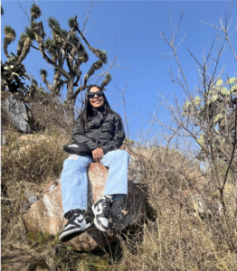 Fotografía de Lizbeth. Ella es una mujer joven que está sentada en una enorme roca. Usa gafas oscuras y luce sonriente.