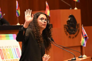 Fotografía de Geras. Elle es una persona que tiene el cabello rizado a la altura del hombro. Se le ve hablando detrás de un micrófono.