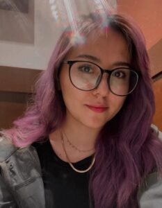 Fotografía de Ana. Ella es una mujer joven de piel blanca que tiene el cabello pintado color morado. Usa lentes de armazón negro y tiene los labios pintados. 