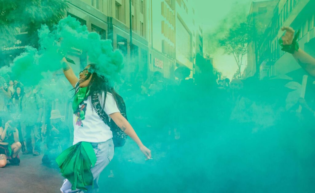 Banner que está ilustrado con la fotgrafía en plano medio de una mujer que porta un pañuelo verde y sostiene con su mano derecha una bengala de humo verde durante una manifestación