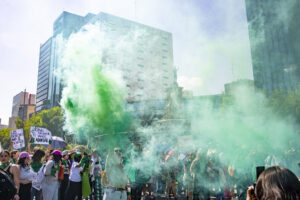 Fotografía en plano general de una manifestación. Está una mujer alzando el brazo derecho sosteniendo una bengala de humo verde.