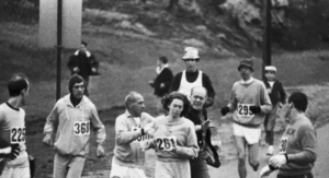 Fotografía en plano general donde se retrata una carrera. Se ve a varios hombres alrededor de Kathrine Switzer, entre ellos al director de carrera intentando sacarla del recorrido.