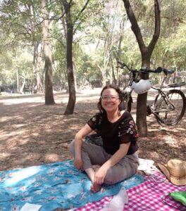 Fotografía de Claudia. Ella es una mujer joven que tiene el cabello a la altura del hombro y que usa lentes. Está sentada sobre una manta en un parque con árboles.