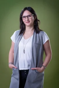 Fotografía en plano americano de Luisa Tello. Es una mujer que tiene el cabello castaño oscuro a la altura del hombro. Viste playera blanca, chaleco gris y pantalón de mezclilla azul. Tiene las manos dentro de los bolsillos del chaleco.
