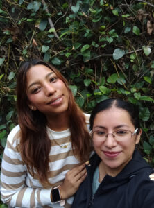 Selfie de Susana y Getzemany. Dos mujeres jóvenes que posan frente a una pared cubierta de hojas. Ambas sonríen.
