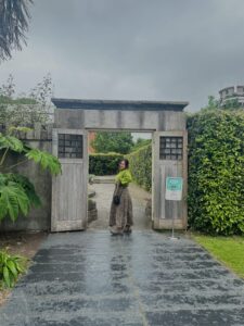 Fotografía de Vane. Ella es una mujer joven que posa sonriente bajo una puerta de madera en un jardín.