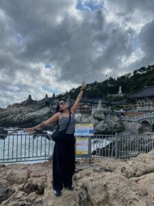 Fotografía de Lizbeth. Ella es una mujer joven que posa sonriente frente a un templo budista que está a orillas del mar