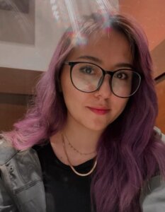 Fotografía de Ana. Ella es una mujer joven de piel blanca que tiene el cabello pintado color morado. Usa lentes de armazón negro y tiene los labios pintados.
