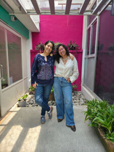 Fotografía de Dunia y Fer. Son dos mujeres que posan sonrientes y abrazadas frente a una pared rosa. 