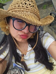 Fotografía de Vanessa. Ella es una mujer joven que tiene los labios pintados de rojo. Usa lentes de armazón negro y sombrero.